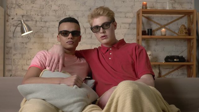 Two gay guys sit on a sofa covered in a warm blanket and watch a 3d scary movie, show an emotion of fright. LGBT lovers, happy gay family, home cosiness concept. Look at the camera 60 fps
