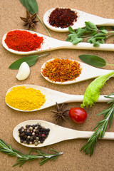 Cooking hot spicy meal. A set of four different spices in wooden spoons, fresh cherry tomatoe, garlic, rosemary and salvia, corkwood background, top view