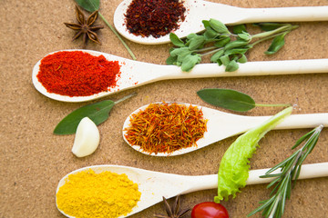 Cooking hot spicy meal. A set of four different spices in wooden spoons, fresh cherry tomatoe, garlic, rosemary and salvia, corkwood background, top view