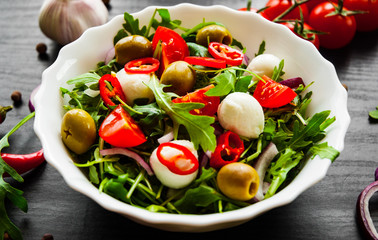 fresh salad with Green rucola, cheese, olive, tomato, onion, pepper in white bowl on dark wooden background