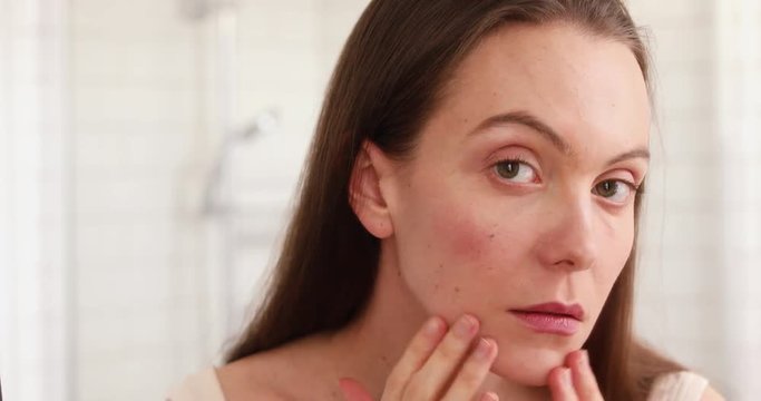 Young beautiful woman touching skin in bathroom. Unhappy girl standinglooking in the mirror, checking dry irritated skin. Morning skincare routine