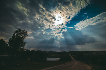 Sun rays are streaming through the clouds. Blue sky and fleece clouds over the summer lake and green trees. Fantastic rural landscape.