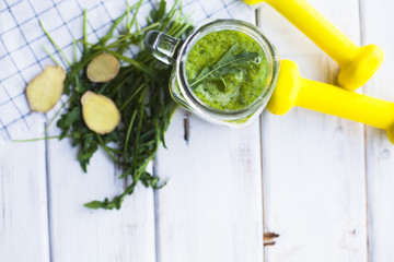 Naklejka premium Healthy Breakfast, Sport, Fitness, Diet Concept. A glass of fruit and vegetable green smoothie, fresh herbs, ginger and yellow dumbbells on a white wooden table with a plaid napkin