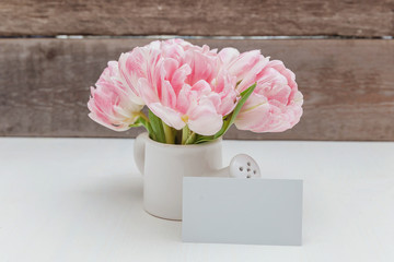 Women's Day March eight. Pink tulip flowers bouquet in small toy watering can vase and greeting card on a wooden background. Post blog social media,view from above, layout mockup for woman day