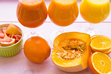 Fototapeta na wymiar Healthy Food, Diet Concept. Three glasses of fruit and vegetable smoothie in a raw with pieces of fresh orange, pumpkin and grapefruit on pink wooden background