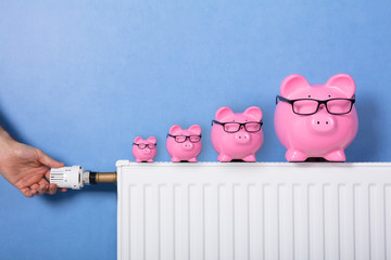 Person's Hand Adjusting Thermostat With Piggy Bank On Radiator