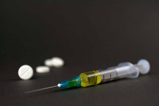 Syringe With A Yellow Drug Lies On A Gray Dark Background, Defocused White Pills, The Concept Of Drug Addiction, Treatment Of Serious Diseases. Selective Focus, Close-up,  Copy-space