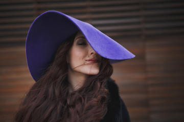 Beautiful smiling charming young woman with long hair in a ultraviolet color hat is on the background of wooden wall. Luxurious brown hair waving from the wind.