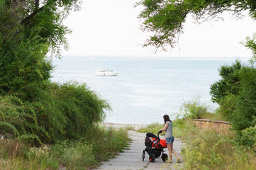 Fototapeta premium Young woman walking at sea background with little cute child baby boy in baby carriage on nature, green trees. Mother, little kid son. Parenthood, family day 15 of may, love, parents, children concept