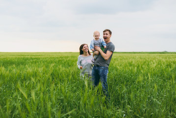 Joyful man, woman walk on green field background, rest, have fun, play, stand with little cute child baby boy. Mother, father, little kid son. Family day 15 of may, love, parents, children concept.