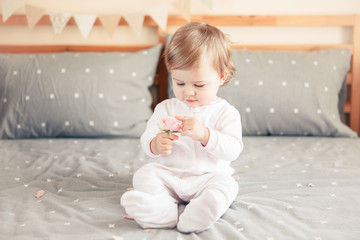 Portrait of cute adorable Caucasian blonde smiling baby girl in white onesie sitting on bed in bedroom and holding pink flower rose. Happy childhood lifestyle concept