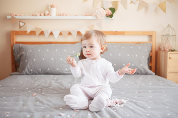 Fototapeta premium Portrait of cute adorable Caucasian blonde smiling baby girl in white onesie sitting on bed in bedroom and holding pink flower rose. Happy childhood lifestyle concept