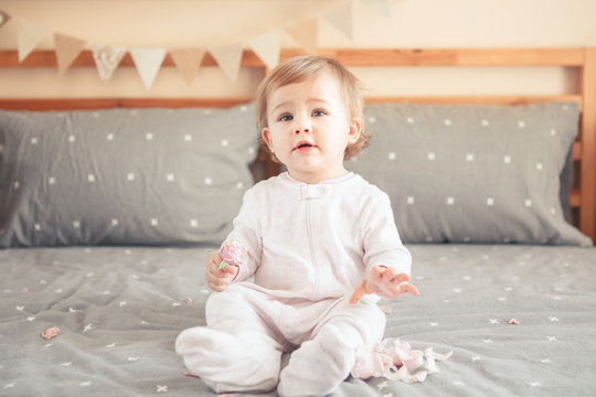 Portrait Of Cute Adorable Caucasian Blonde Smiling Baby Girl In White Onesie Sitting On Bed In Bedroom And Holding Pink Flower Rose. Happy Childhood Lifestyle Concept