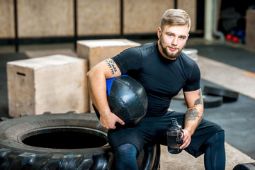 Portrait of a handsome athletic man in black sports wear sitting with ball on the tire in the...