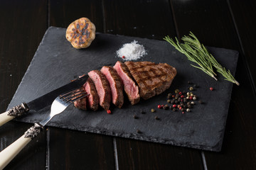 Sliced prime ribeye steak on black stone plate. Medium degree of steak doneness. With rosemary and peppers. Fork and knife.