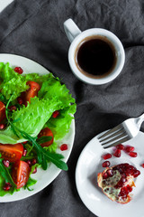Salad on a white plate, espresso, pomegranate and fork on a grey linen napkin. Cherry tomatoes, arugula and pomegranate seeds on fresh lettuce leaves. Flat lay