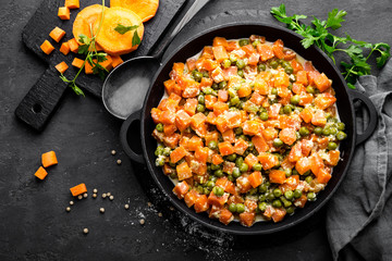 Carrot braised with fresh green peas in creamy milk sauce in stewpan, vegetable saute on black background, top view