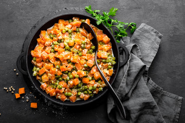 Carrot braised with fresh green peas in creamy milk sauce in stewpan, vegetable saute on black background, top view
