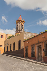 Obraz premium A steep cobblestone street with colorful buildings and a church tower, in San Miguel de Allende