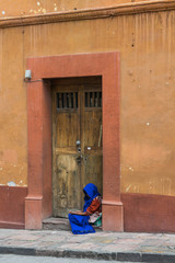A woman sitting in a doorway with old wooden double doors and a colorful old wall, covered in a long blue shawl, in San Miguel de Allende, Mexico