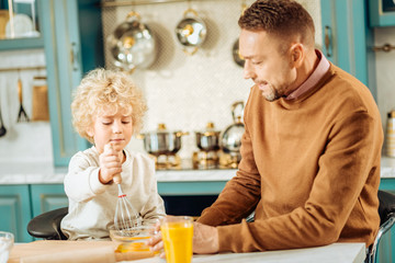 Young cook. Positive nice cute boy sitting at the table and holding whisk while learning to cook