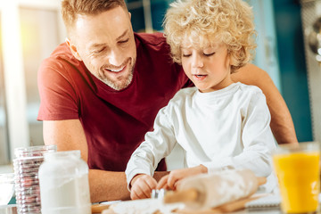 Well done. Handsome nice cheerful man standing near his son and looking at his work while helping his to cook