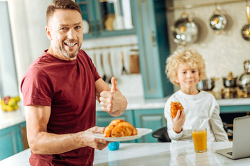 Good job. Happy positive delighted man standing in the kitchen and looking at you while holding a plate with croissants