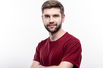 Powerful position. The portrait of a charming young man in a burgundy t-shirt standing half-turned and folding his arms across his chest while posing isolated on a white background