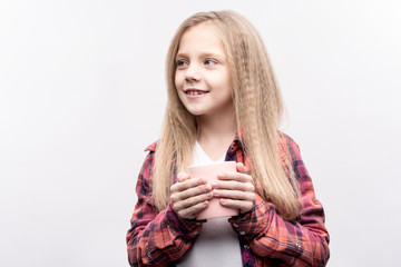 Sweet drink. Cute little girl holding a cup of tea in her both hands and looking away from the camera while posing isolated on a white background