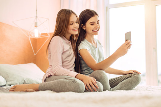 Precious moment. Adorable teenage girls sitting on the bed and taking selfies together while smiling cheerfully at the camera