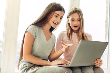 Pleasant discussion. Adorable teenage female friends sitting on the window sill, holding a laptop in their lap and choosing movies for a movie night
