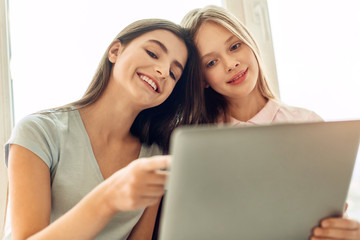 Funny moment. Pleasant upbeat teenage girls sitting on the windowsill and watching a film together while the elder girl pointing at a funny moment and smiling