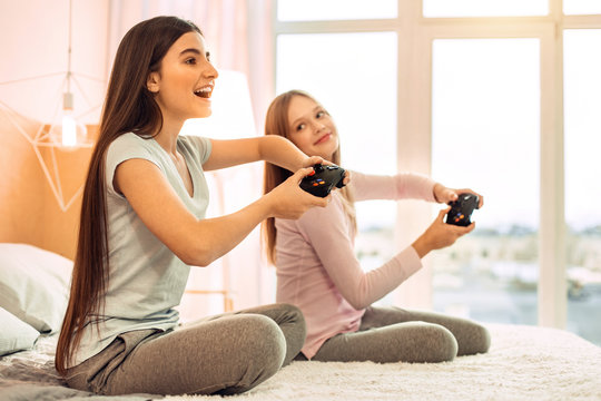 Great pastime together. Charming teenage female friends sitting on the bed and playing video games while one of the girls looking at the other with smile