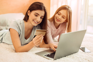 Fast and convenient. Pleasant teenage sisters lying on the bed and doing online shopping together while the older girl paying with her bank card