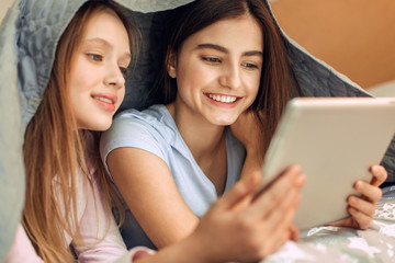 Cozy movie night. Upbeat teenage sisters lying on the bed, hiding under the blanket, and watching a film on the tablet while smiling