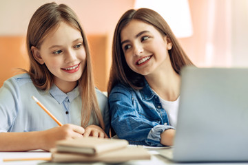 Pleasant supervision. Pleasant teenage girl sitting at the table next to her younger sister and watching her do her home assignment and smiling at her fondly