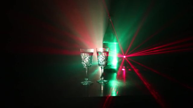 Two glasses of Vodka with bottle on dark foggy club style background with glowing lights (Laser, Stobe) Multi colored. Club drinks theme decoration. Empty space. Selective focus