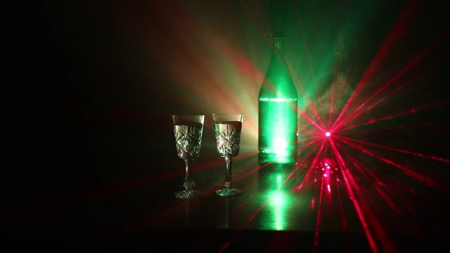 Two glasses of Vodka with bottle on dark foggy club style background with glowing lights (Laser, Stobe) Multi colored. Club drinks theme decoration. Empty space. Selective focus