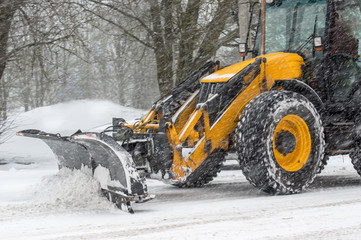 Yellow tractor with snowplow removing snow during heavy snowfall. Winter time street maintenance in...