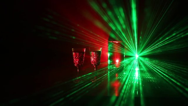 Two glasses of Vodka with bottle on dark foggy club style background with glowing lights (Laser, Stobe) Multi colored. Club drinks theme decoration. Empty space. Selective focus