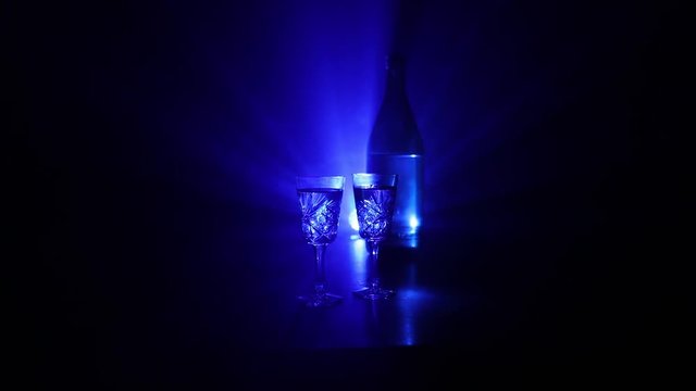 Two glasses of Vodka with bottle on dark foggy club style background with glowing lights (Laser, Stobe) Multi colored. Club drinks theme decoration. Empty space. Selective focus