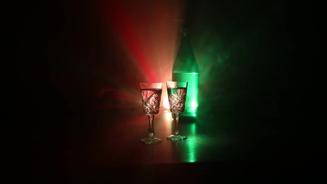 Two glasses of Vodka with bottle on dark foggy club style background with glowing lights (Laser, Stobe) Multi colored. Club drinks theme decoration. Empty space. Selective focus