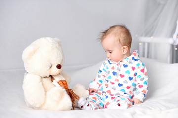 Cute happy baby in a stylish hoodie sitting on a white blanket at the bed and playing with her teddy bear, laughing child.