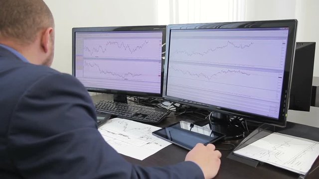 Man financier looks at diagrams on computer screen sitting in office. Specialist watches carefully at images depicted on two monitors, moving mouse with hand. Experienced employee dressed in business