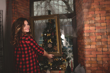 portrait of a beautiful young smiling woman in a shirt with a picture frame on the background of a window and a Christmas tree and a wall of red bricks