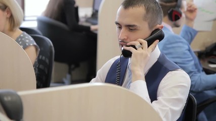 The portrait of the manager who talks on phone with client in the big office among the colleagues. The professional wears dark vast, blue tie and silver fingerring and sit at the table with separation - Powered by Adobe