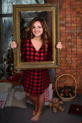 portrait of a beautiful young smiling woman in a shirt with a picture frame on the background of a window and a Christmas tree and a wall of red bricks