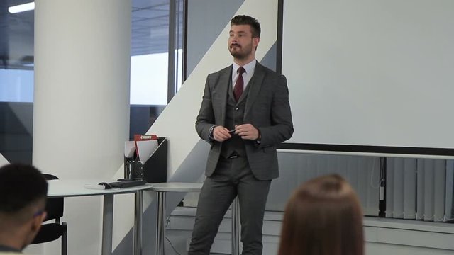 Business trainer conducts training for professionals in large company. Young man walks about room, talks positively and gestures with hands, smiling amicably. Successful coach dressed in business