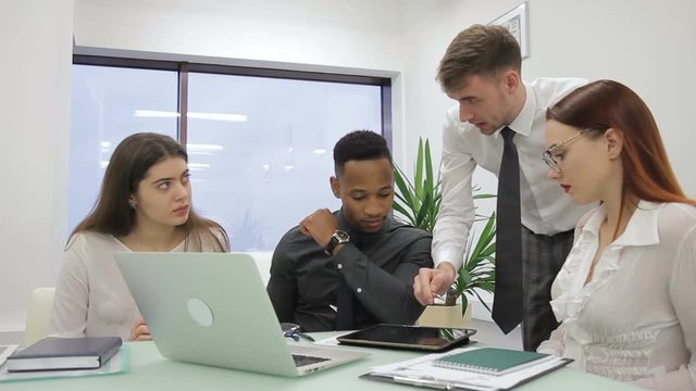 The male boss explains the task to three managers in co-working room. The businessman in tie holds the tablet in his hands and the subordinates listens sitting at the table with laptop, notebook and