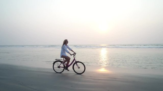 A young pretty girl runs a bicycle that moves along the sandy beach at sunset. The woman spread her hair and they fly in the wind. Aerial view.